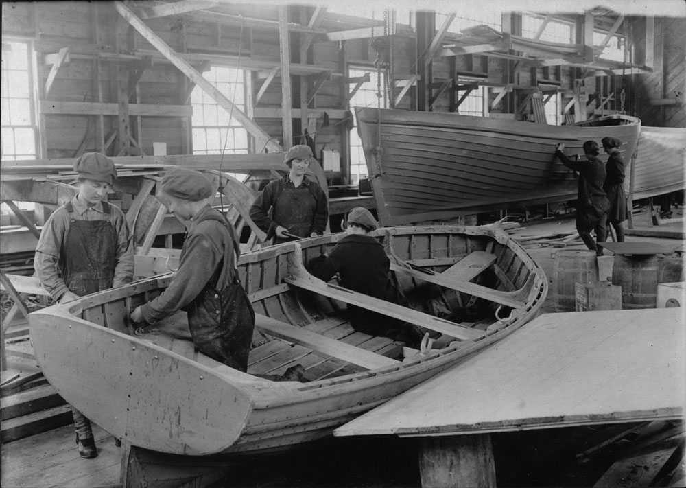 Black and white photograph. Interior of a large wooden building. Women in work clothes, overalls, are performing different tasks constructing  wooden boats.
